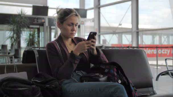 Привлекательная кавказка пользуется мобильным телефоном, сидя в терминале аэропорта. Концепция ожидания рейса, отпуска. Slow Motion — стоковое видео