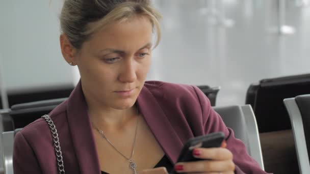 Message de dactylographie féminin caucasien attrayant sur smartphone tout en étant assis à l'aérogare. Réfléchie et concentrée, bouleversée. Le concept d'attendre le vol, les vacances. Mouvement lent — Video