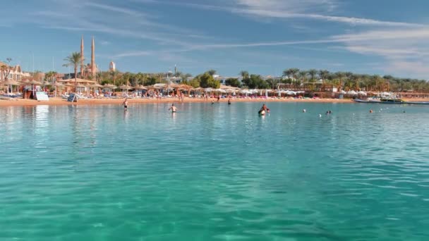 Strand egy trópusi üdülőhelyen, ahol a turisták napfürdőznek és úsznak a tengerben. Gyönyörű türkizkék víz. Szemben a háttérben az esernyők és pálmafák. A távolság egy mecset és a minaret. Lassú — Stock videók