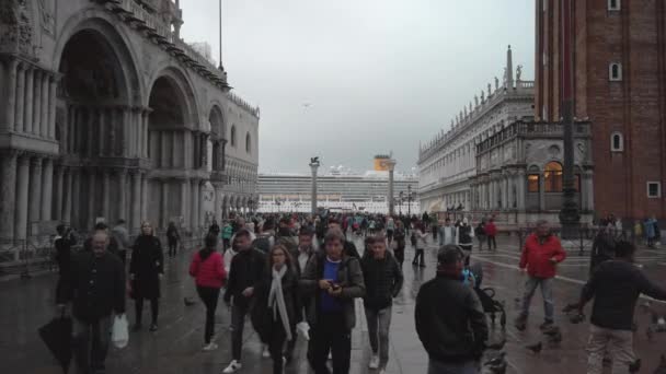Βενετία, Ιταλία-Μάιος 2019: Βενετία, Ιταλία-Μάιος 2019: τεράστια κρουαζιερόπλοιων πλέει στη λιμνοθάλασσα. Οι τουρίστες περπατούν στην πλατεία του Αγίου Μάρκου με θέα τη βιβλιοθήκη της Μαρσιάνα, το παλάτι των Δόγηδων — Αρχείο Βίντεο