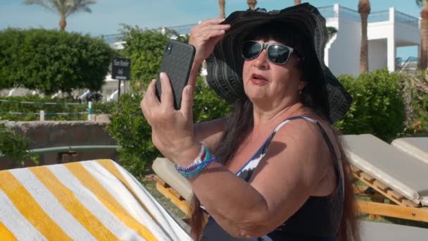 Mujer mayor caucásica de edad avanzada con un sombrero negro hace una selfie en un teléfono inteligente y toma el sol en el sol mientras está sentado en una tumbona. En el contexto de un paisaje tropical, palmeras y — Vídeos de Stock