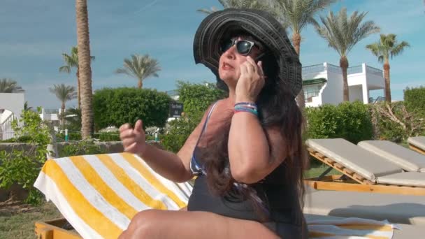 Mujer mayor caucásica de edad avanzada con sombrero negro hablando en un teléfono inteligente y tomando el sol en el sol sentado en una tumbona. Contra el telón de fondo de un paisaje tropical, palmeras y verdes arbustos de verano — Vídeo de stock