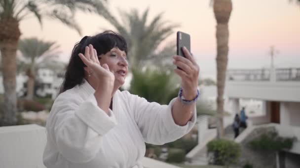 Mulher idosa caucasiana feliz descansando em um resort tropical e fazendo uma chamada de vídeo. Usa um smartphone. Movimento lento — Vídeo de Stock