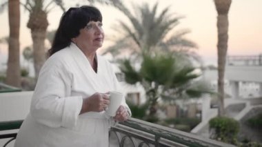 Sabahları otelin terasında kahve içen beyaz yaşlı kadın, tropik bir tatil beldesinde beyaz terry bornozla. Palmiye ağaçlarının ve denizin arka planında. Yavaş Çekim, yakın çekim