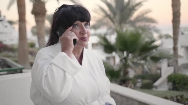 Lächelnde kaukasische Seniorin, die in einem weißen Frottee-Gewand auf einer Hotelterrasse mit einem Smartphone spricht. Zeitlupe, Nahaufnahme — Stockvideo