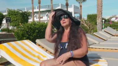 Siyah şapkalı beyaz yaşlı kadın yaşlı, şezlongda otururken güneşte güneşlenenir. Tropik bir manzara, palmiye ağaçları ve yeşil yaz çalılar fon karşı. Yavaş Çekim