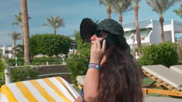 Mujer mayor caucásica de edad avanzada con sombrero negro hablando en un teléfono inteligente y tomando el sol en el sol sentado en una tumbona. Contra el telón de fondo de un paisaje tropical, palmeras y verdes arbustos de verano — Vídeos de Stock