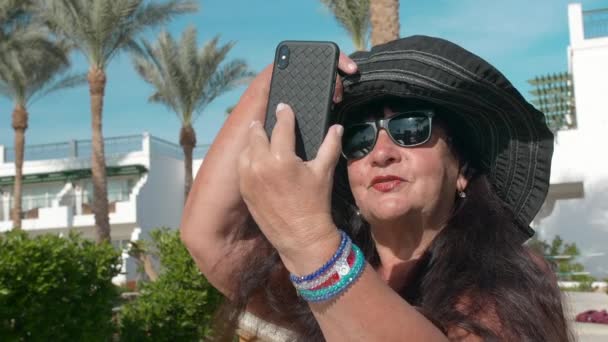 Mujer mayor caucásica anciana en sombrero negro hablando en videollamada en un teléfono inteligente mientras está sentada en una tumbona. Contra el telón de fondo de un paisaje tropical, palmeras y verdes arbustos de verano. El — Vídeos de Stock