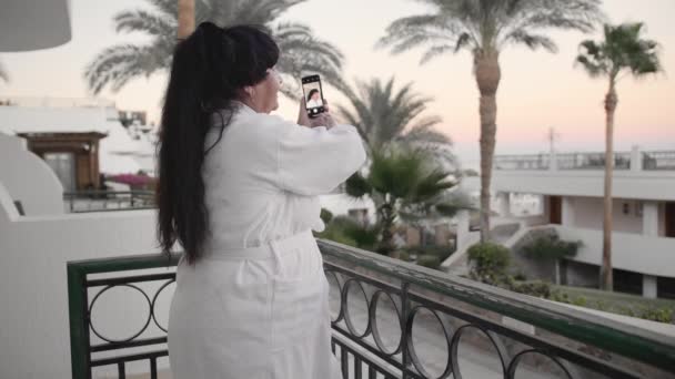 Mulher sênior caucasiana sorridente tira foto selfie no smartphone. Deve estar no terraço do hotel em um manto terry branco em um resort tropical. No contexto das palmeiras e do mar — Vídeo de Stock