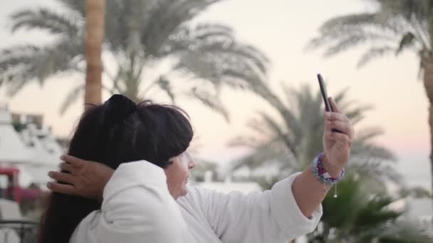 Mujer mayor caucásica feliz haciendo selfie en el teléfono inteligente. Debe estar en la terraza del hotel con una bata de rizo blanco en un resort tropical. Contra el telón de fondo de palmeras y el mar. El — Vídeo de stock