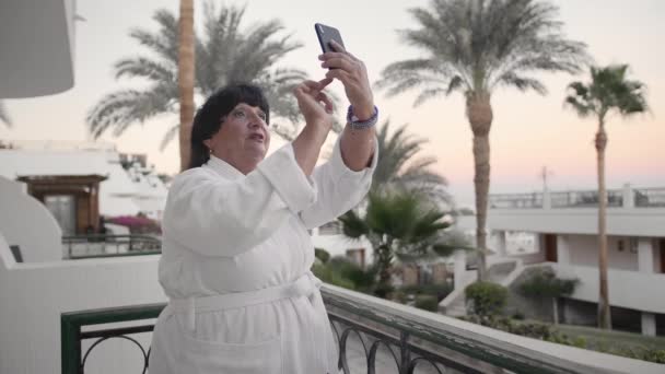 Beyaz yaşlı kadın akıllı telefonda fotoğraf çekiyor. Otelin terasında, tropik bir tesiste beyaz terry bornozla olmalıdır. Palmiye ağaçlarının ve denizin arka planında. Kavram — Stok video
