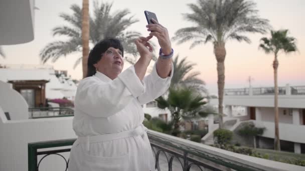 Donna anziana caucasica scatta foto su uno smartphone. Dovrebbe essere sulla terrazza dell'hotel con un accappatoio di spugna bianca in un resort tropicale. Sullo sfondo delle palme e del mare. Il concetto — Video Stock