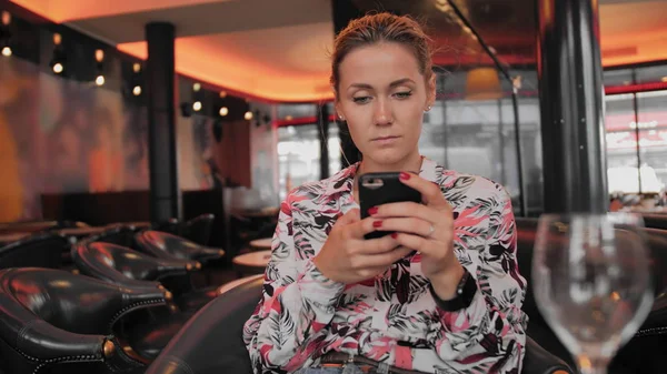 Une jeune Française utilise les réseaux sociaux pour communiquer avec un téléphone portable. Assis dans un restaurant sur une chaise en cuir attendant votre commande. Au premier plan des lunettes floues. Slow Motion Dolly Shot Photos De Stock Libres De Droits