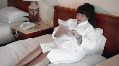 beyaz terry bornoz bir otelde bir yatakta yatan olgun kadın bir tablet kullanır, internet sitelerinde sörf. Yavaş Çekim