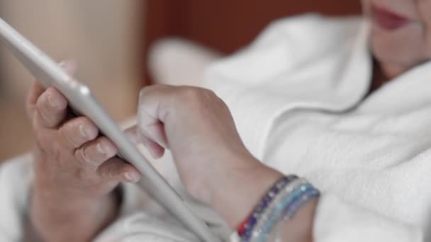 Пожилая женщина, лежащая на кровати в белом махровом халате, пользуется планшетом, просматривает интернет-сайты. Медленное движение, крупный план — стоковое видео