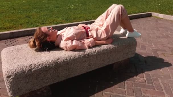 Stanca, ragazza caucasica giace appoggiata su una panchina di pietra in una chiara giornata di sole. Il concetto di stanchezza e relax, duro lavoro — Video Stock