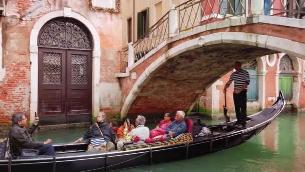 VENECIA, Italia - Mayo 2019: Los turistas multirraciales viajan en góndola a través de los estrechos canales de Venecia. Utilice los teléfonos inteligentes para tomar fotos. Navega bajo un pequeño puente. Movimiento lento — Vídeo de stock