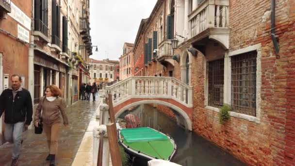 Wenecja, Włochy-2019 maja: Jesienna deszczowa pogoda w mieście. Ludzie chodzą wzdłuż wąskiej ulicy obok kanału, na którym łódź jest zacumowany. Mały most jest typowym wejściem do budynku — Wideo stockowe