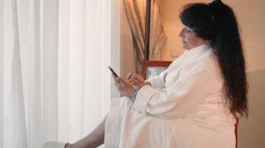 mutlu, gülümseyen Kafkas yaşlı kadın beyaz bir bornoz büyük bir panoramik pencere yakınında otururken bir mesaj yazarak, bir akıllı telefon kullanıyor. Yaşlı insanlar tarafından gadget'lar kullanma kavramı. Yavaş
