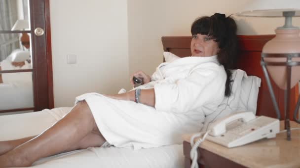 Kaukasiska kvinnan ligger på en säng i ett hotell i en vit badrock och styr TV med fjärrkontrollen. Konceptet att använda teknik och prylar av mogna människor i ålder. Slow motion — Stockvideo