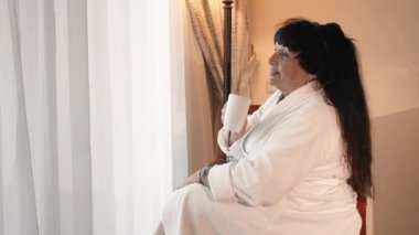 mutlu, gülümseyen Kafkas yaşlı kadın beyaz bir terry bornoz büyük bir panoramik pencere yakınında sabah kahve içiyor. Yavaş Çekim