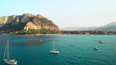 Palermo, Sicilya, İtalya - Ağustos 2019: Kayalık bir dağın, adaların ve turkuaz denizin arka planında beyaz yelkenli lüks yatlar. Havadan insansız hava aracı vuruldu