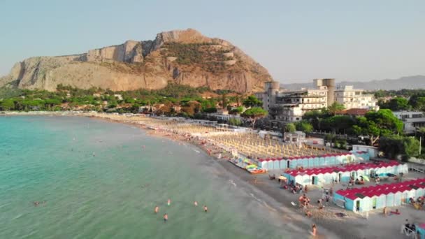 パレルモ、シチリア、イタリア:カメラは海の海岸線の上をスライドします。ビーチで多くの旅行者や観光客。太陽から対称的に配置されたパラソル。背景に大きな岩。空中ドローンショット. — ストック動画