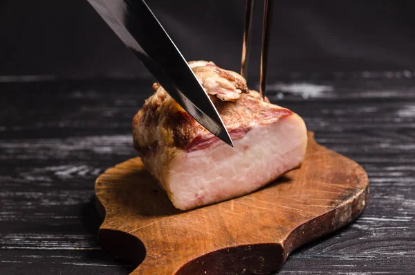 Meat carving using a carving knife and carving fork. A piece of smoked bacon, carving on a wooden cutting board.