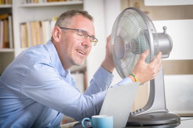 Adam ofiste çalışırken sıcaktan muzdarip oluyor ve vantilatörün karşısında serinlemeye çalışıyor.