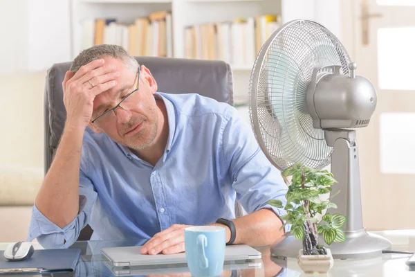 Man suffers from heat while working in the office and tries to cool off by the fan