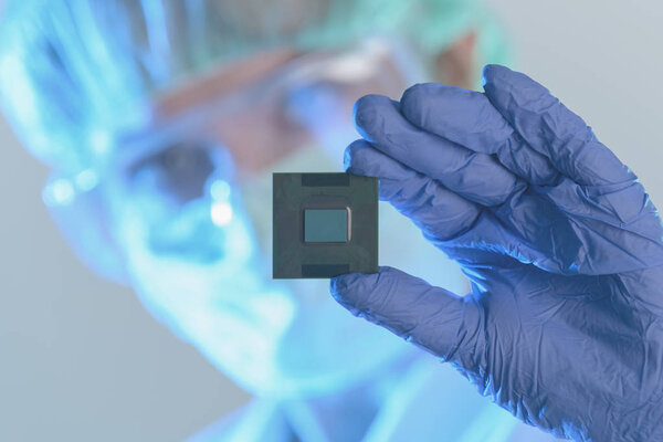 An engineer working in a laboratory wearing a special uniform and protective gloves holds new processor in hands and examines it