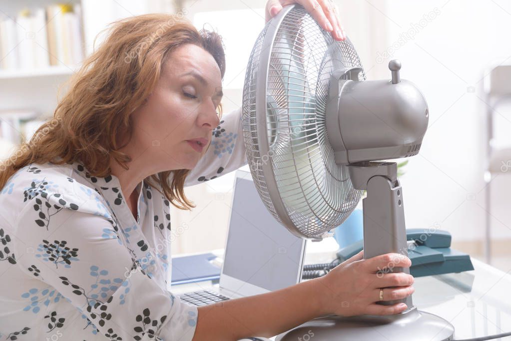 Woman suffers from heat while working in the office and tries to cool off by the fan