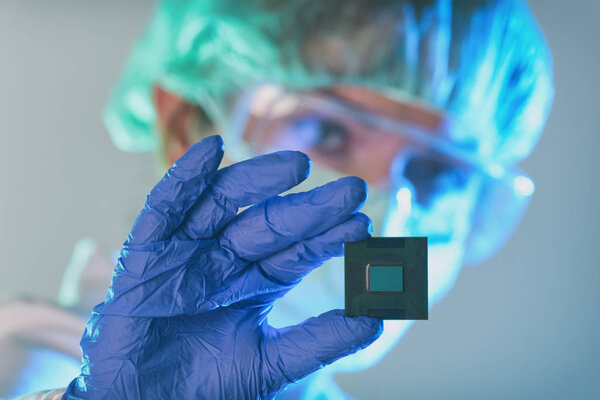 An engineer working in a laboratory wearing a special uniform and protective gloves holds new processor in hands and examines it