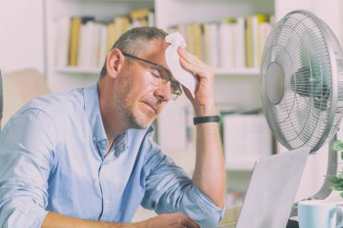Man suffers from heat while working in the office and tries to cool off by the fan clipart