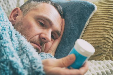 Depresyondan muzdarip yetişkin bir adam yatağında bir battaniyeyle uzanıyor ve elinde bir şişe antidepresan tutuyor.