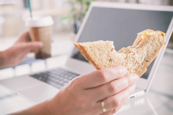 Frau isst ein Frühstückssandwich, während sie mit einem Laptop arbeitet — Stockfoto