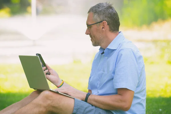 男人坐在公园的草地上 拿着笔记本电脑 用手机聊天 — 图库照片