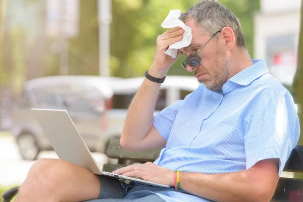 Man suffers from heat while working with laptop outside and wiping his forehead with tissue