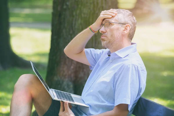男人在公园里一边用笔记本电脑一边擦拭额头 一边忍受着热量的折磨 — 图库照片