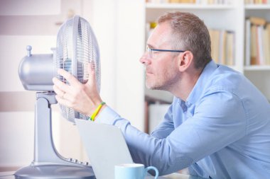 Adam ofiste çalışırken sıcaktan muzdarip oluyor ve vantilatörün karşısında serinlemeye çalışıyor.