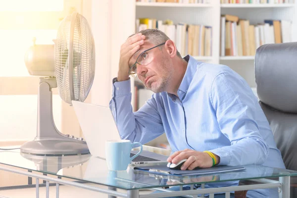 男はオフィスで働いている間に熱に苦しみ ファンによって冷却しようとします — ストック写真