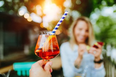 Hayatı kutluyoruz. Aperol spreyli tost. Arka planda gülümseyen genç bir kadın. İnce film tahıl dokusu.