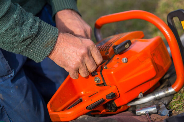 Old man in blue pants repair orange chainsaw placed on the ground with his bare hands.