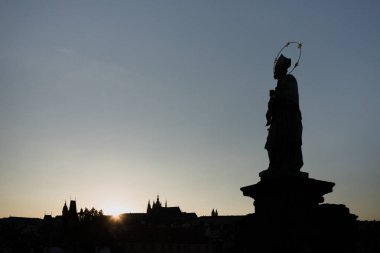 Prag'daki Charles Köprüsü'ndeki Nepomuk Heykeli'nden Aziz John'un silueti, gün batımında arka planda St. Vitus Katedrali ile.