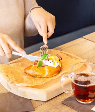 Kadın bir restoranda kahvaltı yapıyor, yumurta kremalı kruvasan ile ahşap bir tabakta yemek yapıyor, brunch tarzı yemek yiyor.