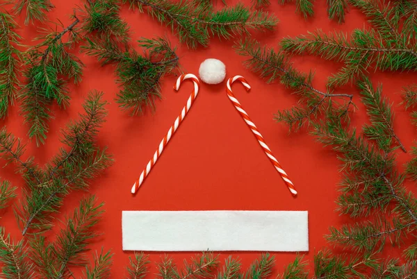 Criativa de chapéu de Papai Noel, conceito mínimo de Natal. Bengalas doces, pompom, guarnição em fundo vermelho com ramos de pinho — Fotografia de Stock