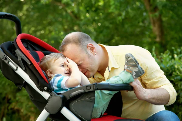 Utomhus porträtt av en lycklig far och son. baby sitter i barnvagnen och pappa i sommar park — Stockfoto