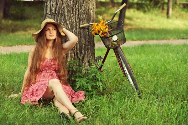 Jovem mulher de vestido e chapéu com bicicleta sentada em um parque perto de uma árvore — Fotografia de Stock