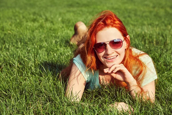 Relaxační zrzka dívka v sluneční brýle ležící na trávě. žena relaxační venkovní. — Stock fotografie