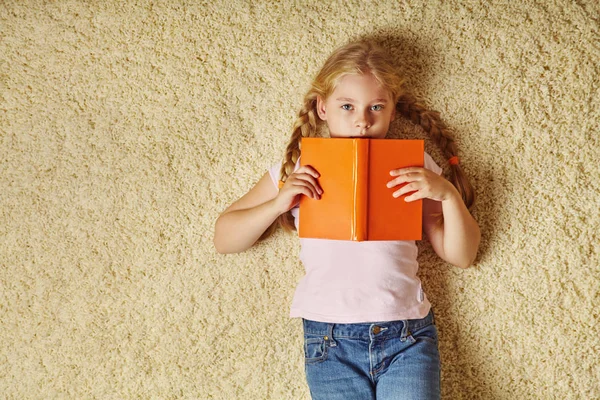 Dívka školního věku ležící na koberci s výhledem na knížku. dítě doma. — Stock fotografie
